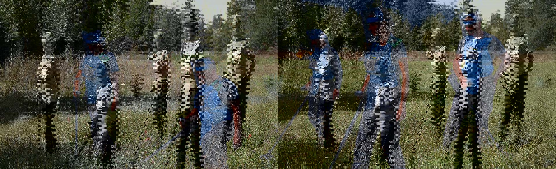 Демінери в захисному спорядженні з детекторами проводять розмінування у високій траві на фоні стіни дерев