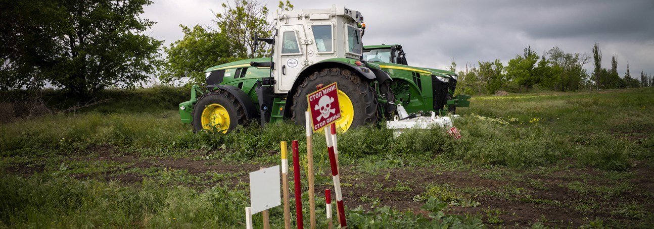 A large agricultural machine stands in the middle of a field behind a landmine warning sign.