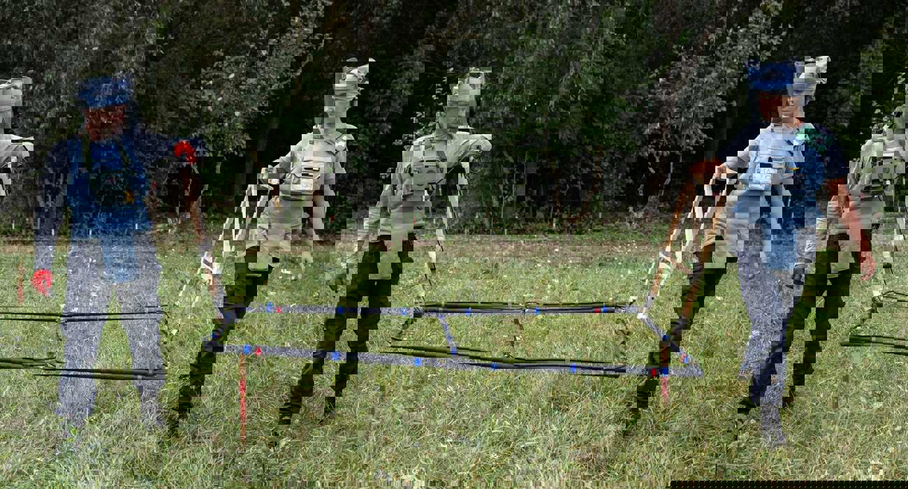 Двоє демінерів тримають над земелю широкоформатний рамковий детектор під час розмінування