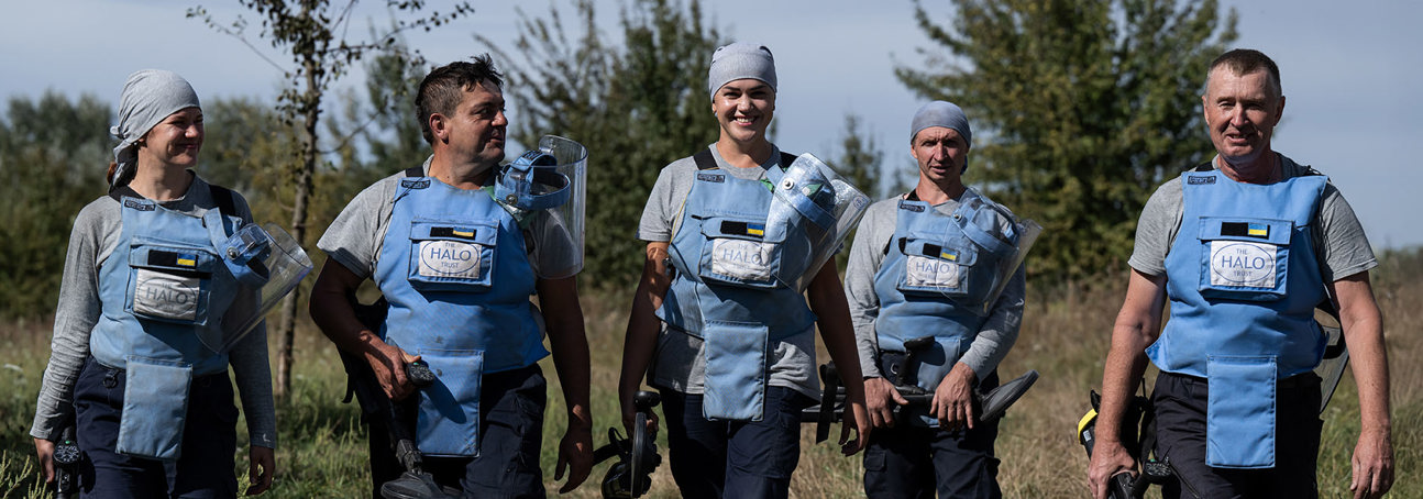 Група демінерів в захисному спорядженні з детекторами, посміхаючись, йде по полю на фоні трави та подиноких дерев