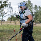 Жінка в захисному спорядженні з детектором проводить розмінування на фоні знака мінної небезпеки