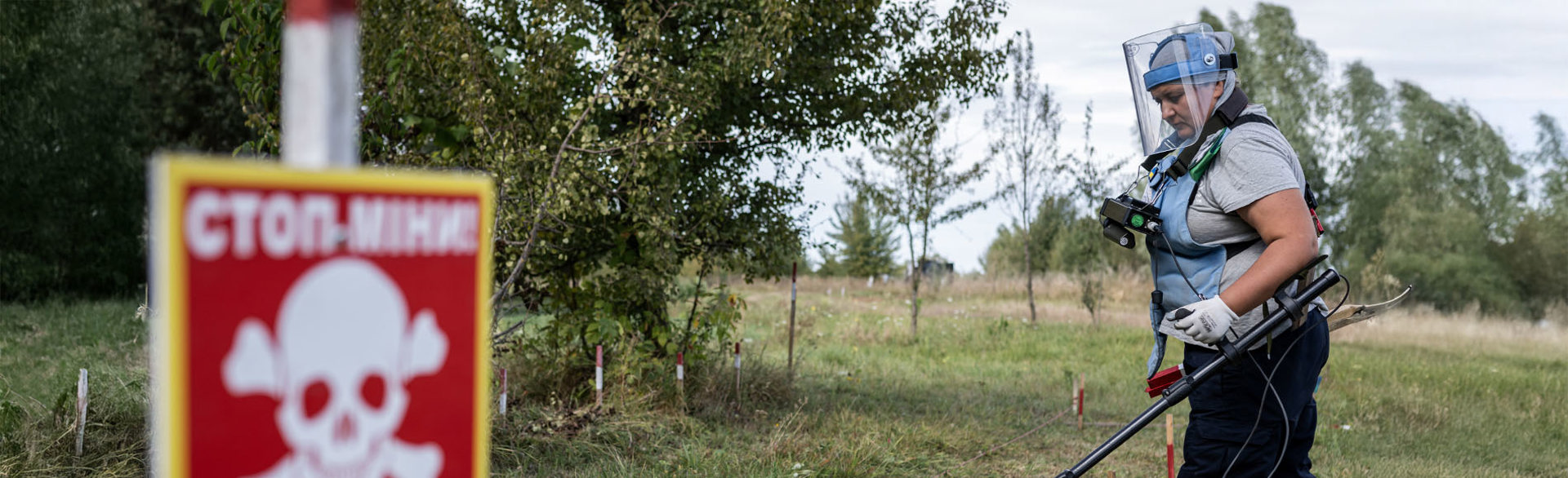 Жінка в захисному спорядженні з детектором проводить розмінування на фоні знака мінної небезпеки