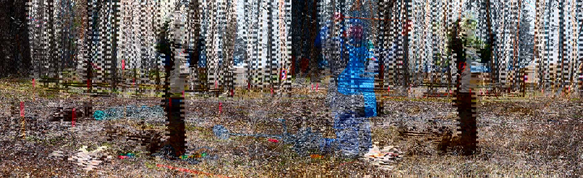 Демінер в захисному спорядженні з щупом в руках проводить пошук розтяжок на фоні інструментів та детектора в осінньому лісі