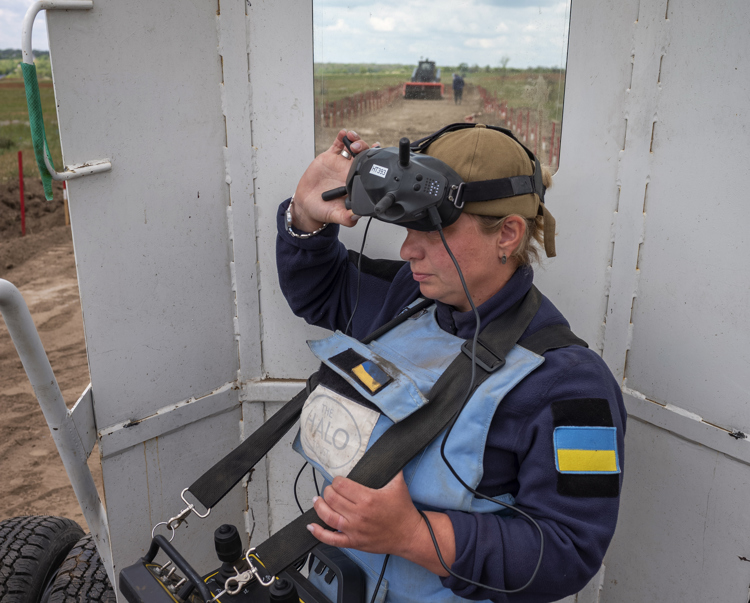 A woman in a HALO Trust vest operates a large remote control and looks into a pair of goggles.