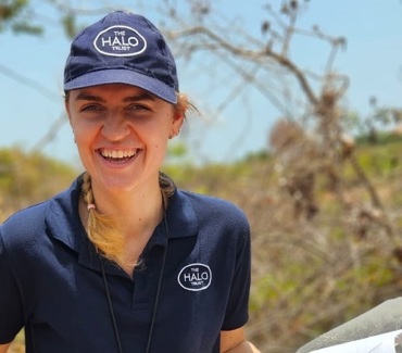 Портрет усміхненої експертки з розмінування в синій футболці і в кепці з пристроєм GPS на шиї і документами в руках на фоні висушеного спекою пейзажа