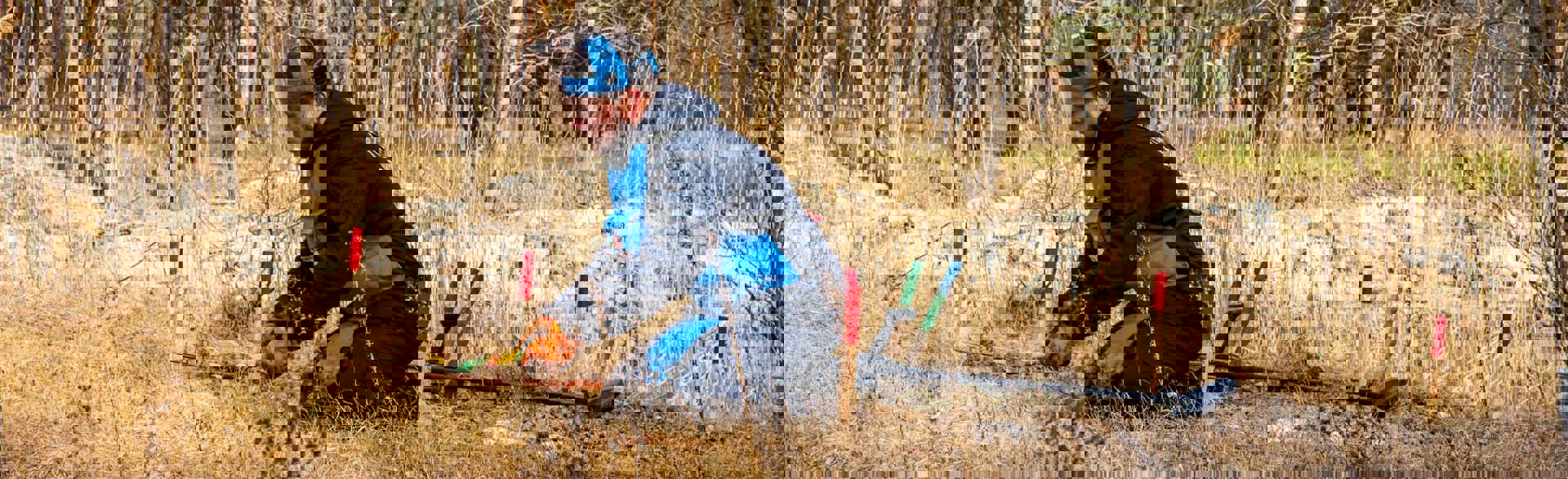 Демінер в захисному спорядженні зрізає зайву рослинність під час розмінування на фоні осіннього лісу