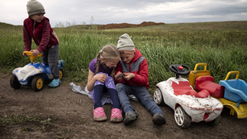 Two young children play together in a field. One boy rides a toy car to the left side.