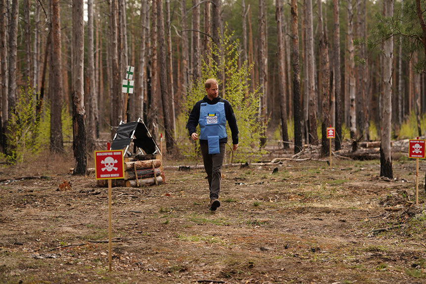 Prince Harry, wearing a HALO vest with the Ukrainian flag, walks through a wooded area featuring a "danger: mines" sign.