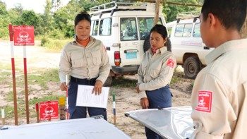 Task supervisor, Ly Sreyneang and unit commander, Pat Liv, stand behind tables with mine warning signs behind them