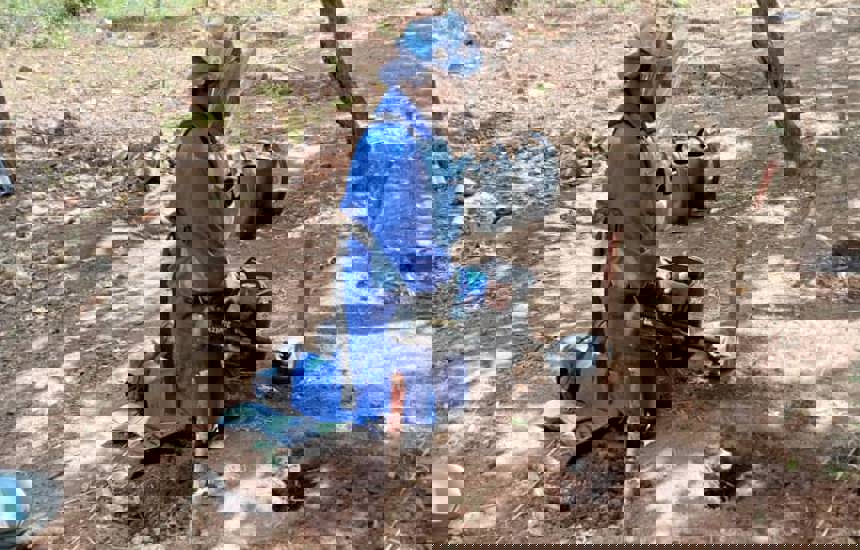 GIS specialist, Delight, wears PPE and holds a metal detector whilst kneeling to find mines in the ground 