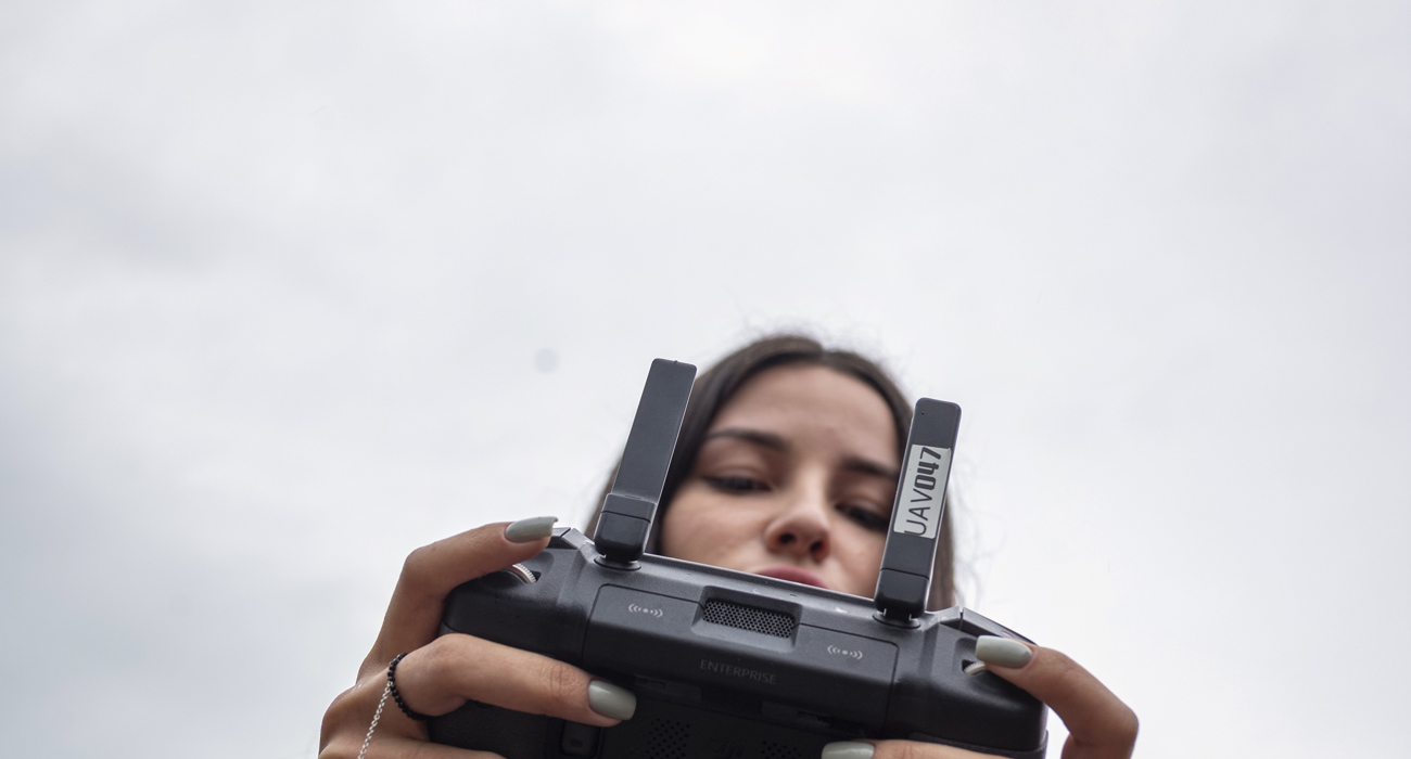 A woman in a blue jacket holds a controller while a drone flies overhead.