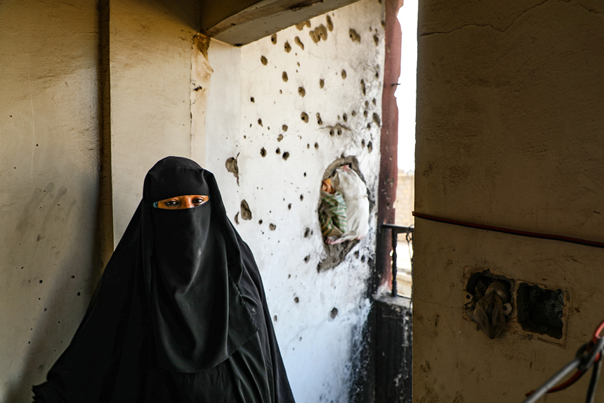 A woman in a niqab veil standing in front of a wall damaged by gunfire and a rocket strike