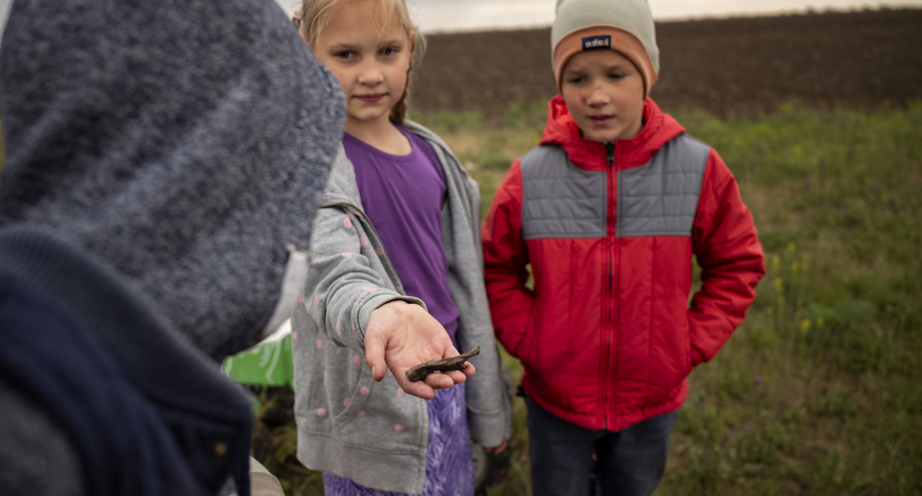 Three young children play together in a field. A girl stands in the middle, showing a small brown object to the camera.