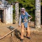 A deminer in PPE uses a rake on the soil in front of a destroyed building