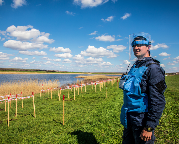 A HALO Trust deminer stands in a rural minefield next to a lake, with contamination clearly marked in front of him