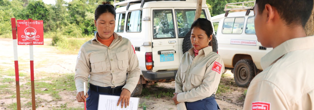 Task supervisor, Ly Sreyneang and unit commander, Pat Liv, stand behind tables with mine warning signs behind them